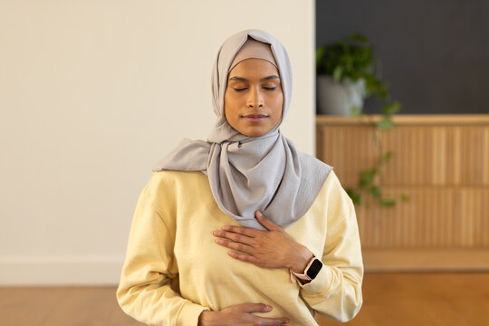 Woman practicing breathing exercise sitting on hardwood floor at home wearing hijab and smartwatch