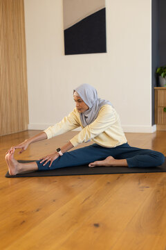 Adult African woman doing seated stretch on black mat at home, wearing headscarf and smartwatch