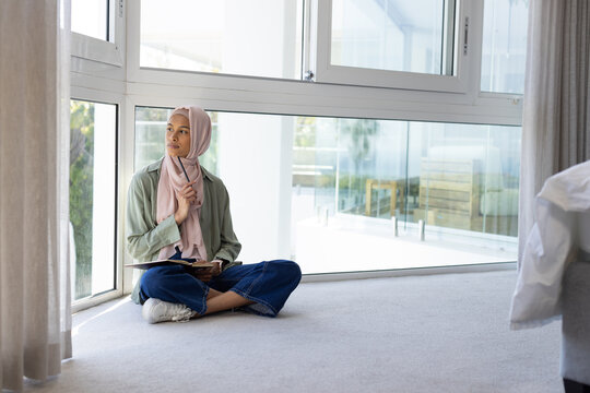 African American woman sitting on carpet by window wearing pink hijab holding notebook, copy space