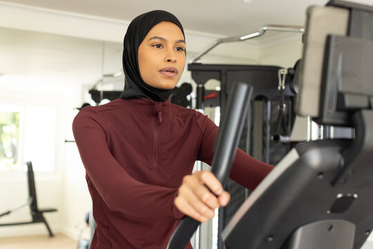 Woman exercising on elliptical at gym wearing black headscarf and maroon zip-top using console