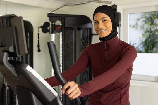 Woman wearing black sports hijab, maroon top using elliptical in compact gym near weights