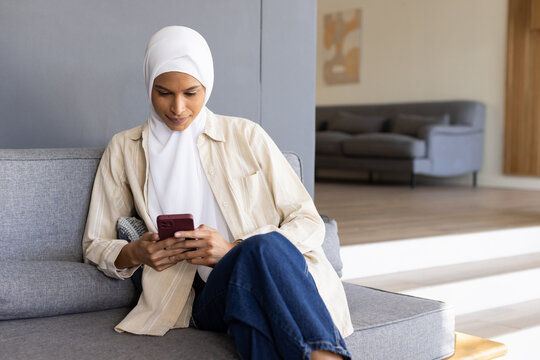 Woman wearing white hijab sitting on grey couch in living room holding maroon phone and scrolling