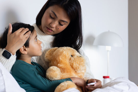 Asian mother and son sitting on bed, mother checking temperature while son cradling teddy bear