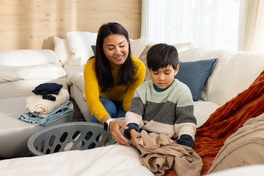 Asian mother in mustard top and school-age son folding beige garment at home with laundry basket