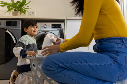 Diverse mother in mustard top and school-age son passing knit garment into white basket by washer