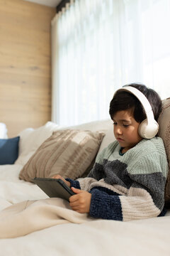 Asian child wearing sweater sitting on sofa using tablet listening with white headphones