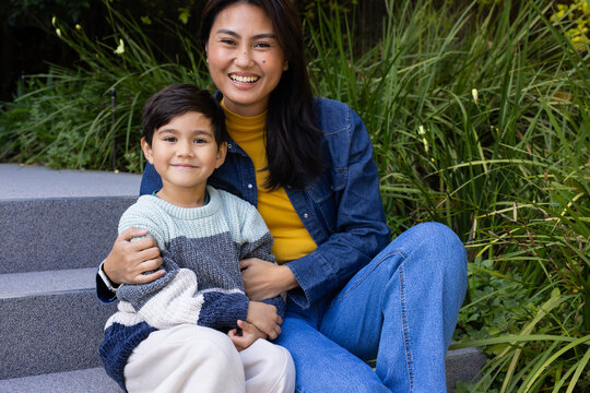 Diverse mother and child sitting on concrete steps by tall grasses, in denim jacket, chunky sweater
