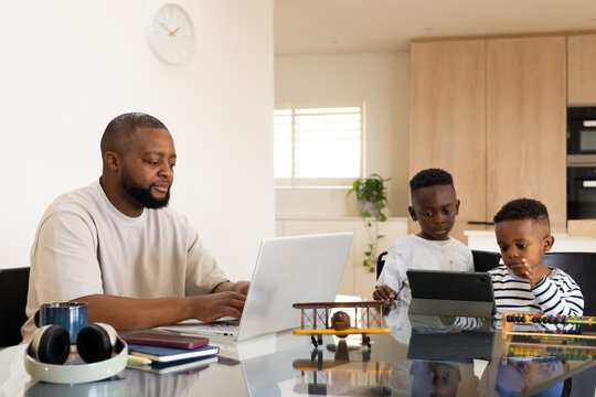 Father and two boys sitting at glossy kitchen table, working on laptop, using tablet and abacus