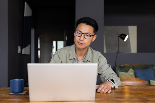 Mid adult Asian man sitting at wooden table using silver laptop wearing green shirt and eyeglasses