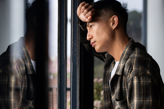 Mid adult Asian man leaning on window in plaid flannel, white tee, gazing outward with reflection