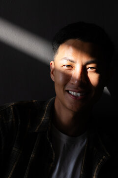 Mid-adult Asian man smiling under diagonal beam in room, shadow, plaid shirt, white tee, copy space