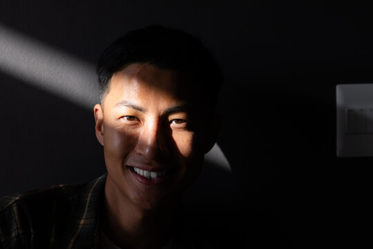 Asian mid-adult man smiling, facing camera in dim room with diagonal light beam and switch panel