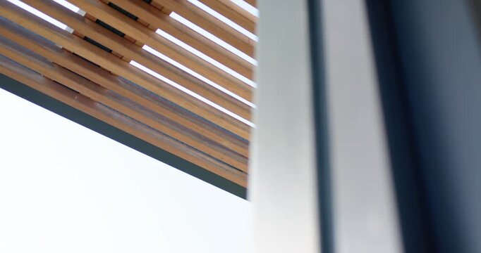 Camera is shifting left, tilting upward, exposing slatted canopy on facade, pale sky, showing form
