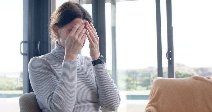 Adult woman sitting on chair pressing palms over face, lowering hands, wiping tears by glass doors