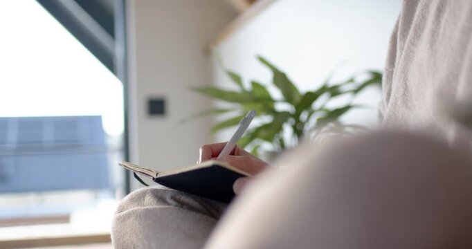 Woman is placing pen tip and writing in black notebook on lap near window for journaling