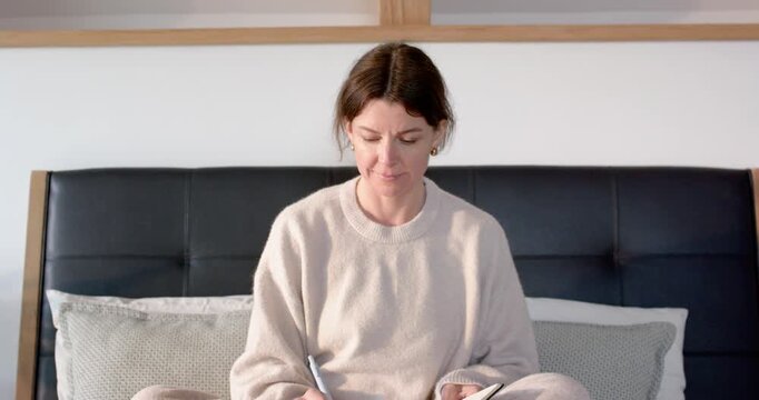 Adult woman holding black notebook and pen in beige loungewear, writing on bed while glancing right