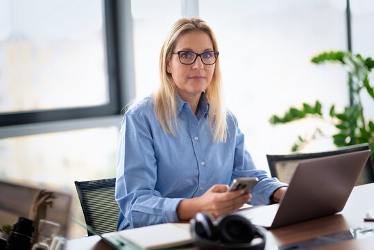 Multitasking businesswoman holding a smartphone while working on a laptop in a modern office