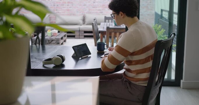 African American man viewing tablet, tapping screen, writing notes while studying at dining table