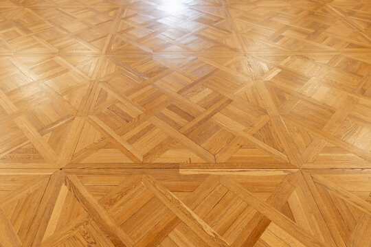 Parquet floor showcases an intricate geometric pattern in polished wood, with visible grain, warm amber tones, and subtle light reflections. The clean indoor surface provides an elegant background