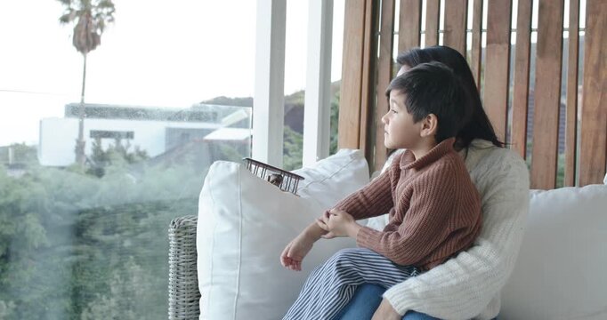 Diverse mother and child son sitting on white sofa by glass railing, boy pointing, mother guiding