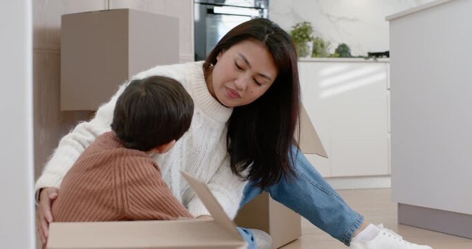 Diverse mother and son kneeling after moving boxes unpacking toys on kitchen floor