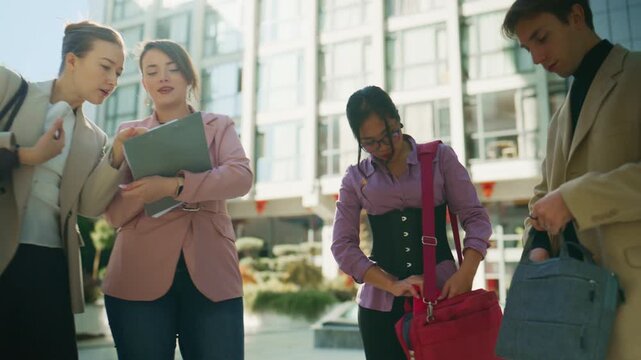 Young professionals engage in an outdoor business meeting, exchanging documents and discussing ideas. A diverse team collaborates on a project outside a modern corporate building.