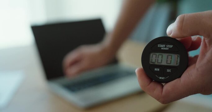 Close up of a man holding a digital stopwatch while starting a laptop to measure how long the computer takes to boot