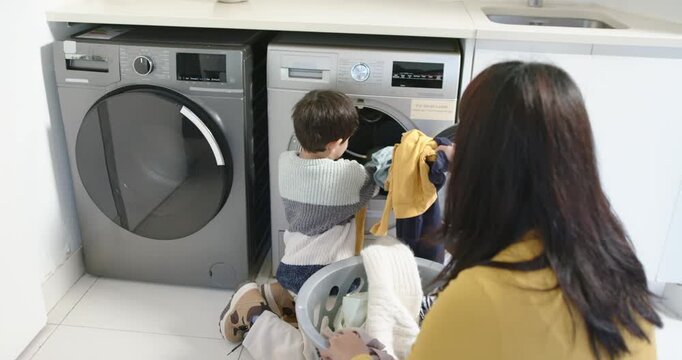 Diverse mother and son kneeling at laundry with washer door open, sorting clothes into basket