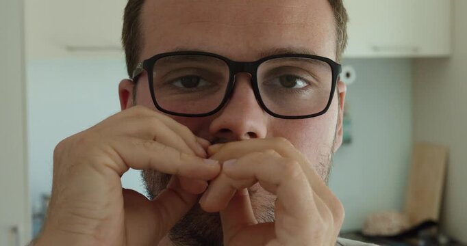 A man wearing glasses places a small snus pouch beneath his lower lip using both hands indoors