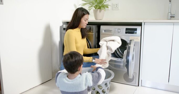 Asian mother and son passing clothes from basket in laundry room, loading washer to wash clothes