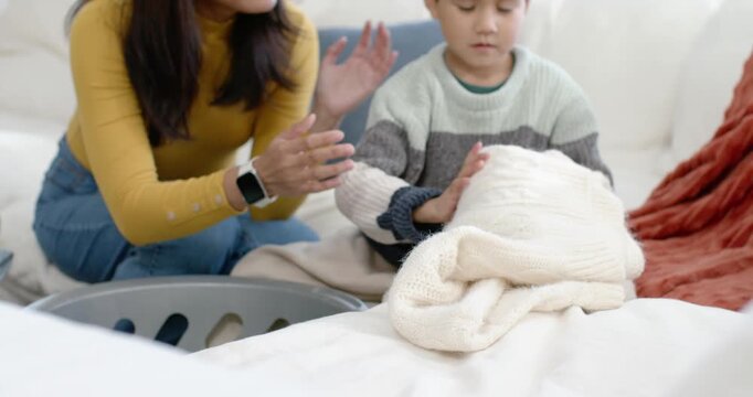 Asian woman wearing mustard guiding child folding blanket on couch, teaching folding near gray bin