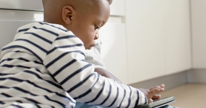 African boys on kitchen floor sharing tablet, younger tapping screen while playing