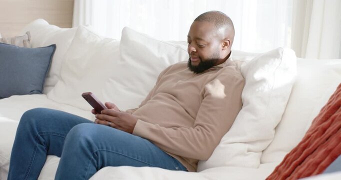 African American man sitting on sofa at home holding phone, tapping after opening message reacting