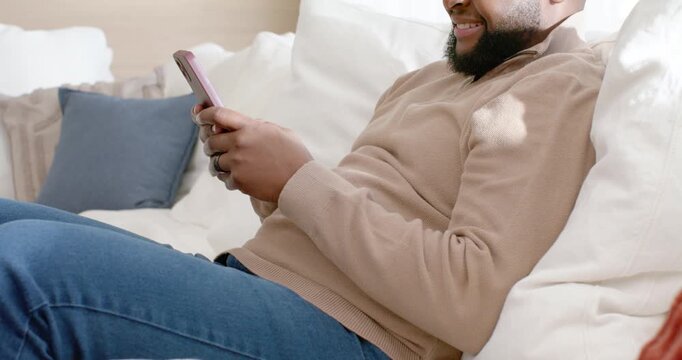 African-American man reading ping lying on sofa in tan-knit tapping pink cell smiling, copy space