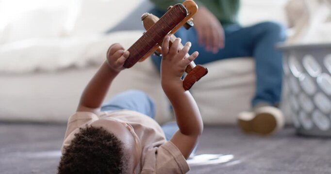 African American toddler turning wooden plane to camera, father folding from laundry basket on sofa