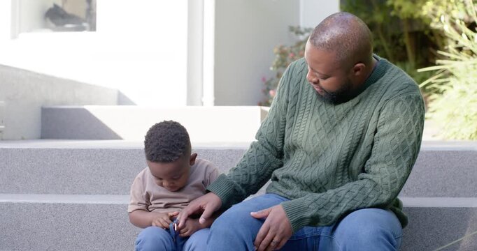 African dad and child sitting on front steps, child fidgeting with toy, dad guiding hands