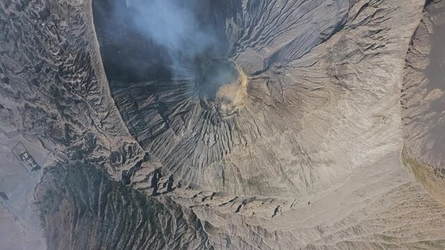 Topdown drone fisheye shot of Mount Bromo crater emitting smoke over rugged volcanic landscape.