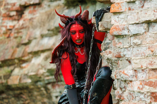 Demonic cosplay woman with horns and chains posing on old brick wall