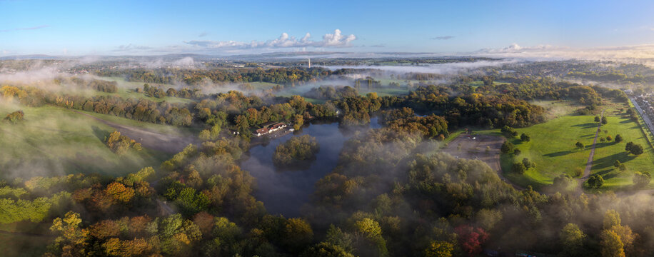 Aerial panorama of Heaton Park Boating Lake and autumn woodland with wisps of morning mist, BT Tower and hills beyond, Manchester