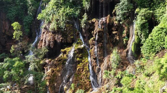 Ground level view of thin cascading waterfalls over mossy limestone cliffs amid lush tropical forest cave entrance.