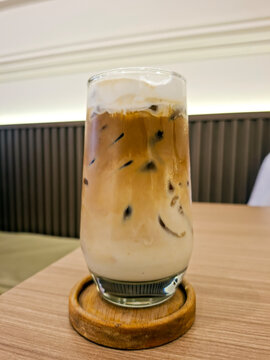 Side view of a layered iced coffee drink topped with thick cream, served in a glass on a wooden coaster inside a cozy cafe.