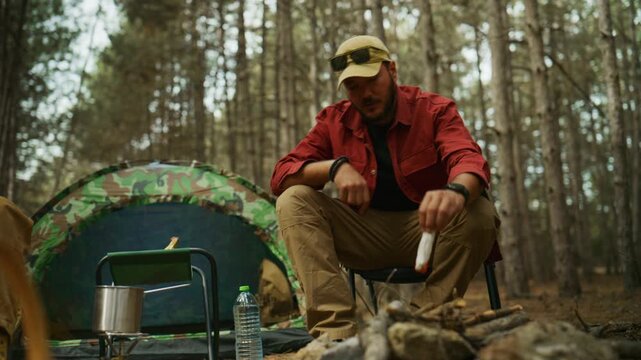 Camper unwraps a fire starter and places it in a campfire ring while camping at a pine forest campsite beside a tent.