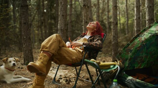 Couple relaxes at a forest campsite with their dog as the man sits down and adjusts a camping stove.