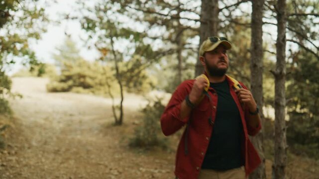 Male hiker walks along a dirt trail in a pine forest, adjusts his backpack straps, and continues deeper into the woods.