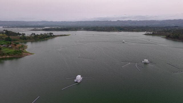Aerial drone panorama of Karangkates dam reservoir with floating intake towers, shoreline farms and distant misty mountains.