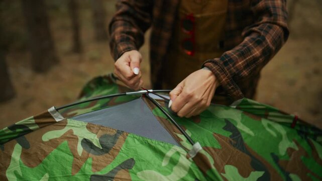 Red-haired woman sets up a camouflage camping tent, ties the top pole knot, then walks into the pine forest.