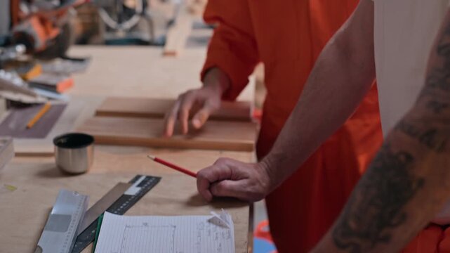 Middle aged Caucasian carpenter and senior craftsman discussing blueprint on workbench in prison woodworking workshop. Perfect for vocational education, trade apprenticeship and craftsmanship