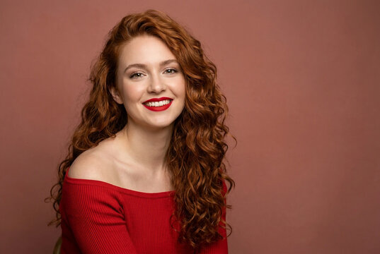 Mujer joven y atractiva con cabello pelirrojo rizado y abundante, sonriendo felizmente. Su&eacute;ter rojo ajustado de manga larga sobre un tono terracota c&aacute;lido, moda de oto&ntilde;o, bienestar femenino, belleza