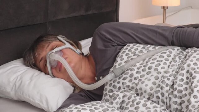 Middle-aged woman with graying hair sleeping on her side wearing a CPAP mask, lightly snoring and adjusting the mask on her face during the night.