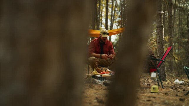 Couple camping in a pine forest feeds their dog a treat while relaxing beside a fire pit and hammock.
